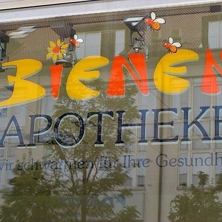 Schaufenster mit dem Text 'BIENEN APOTHEKE' und Cartoon-Bienen, die eine Gebäudefassade widerspiegeln.