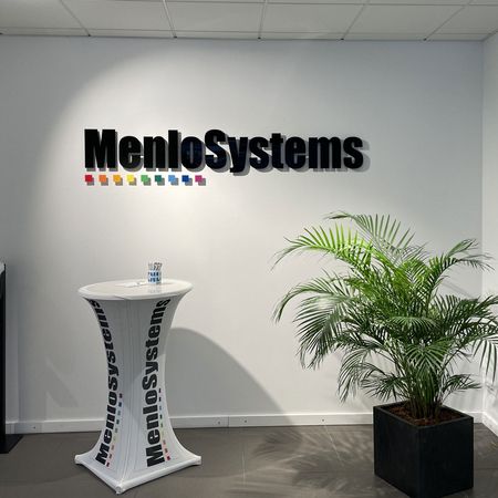 Büroraum mit dem 'MenloSystems'-Logo an der Wand, einem Prospektständer, einem Desinfektionsmittelspender und einer Topfpflanze.