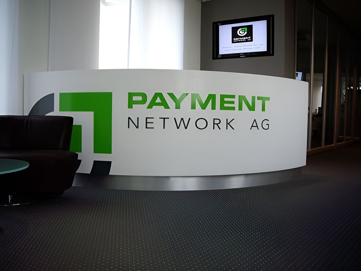 Empfangsbereich mit einem 'PAYMENT NETWORK AG'-Schild auf dem Schreibtisch und einem Firmenlogo auf einem Monitor im Hintergrund.